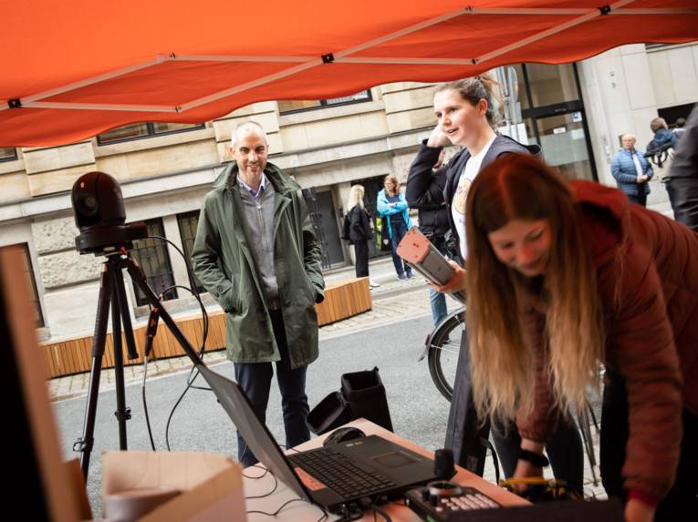 Mann vor einem Pavillon. Darin zwei junge Menschen mit Monitoren.